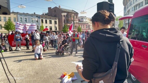 Mehrere Menschen halten Fahnen und Plakate hoch. Im Vordergrund eine Sprecherin der Kundgebung. © Screenshot 